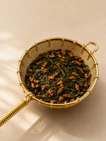 Close-up of a gold mesh tea strainer filled with genmaicha tea against a soft neutral beige background with warm natural shadows