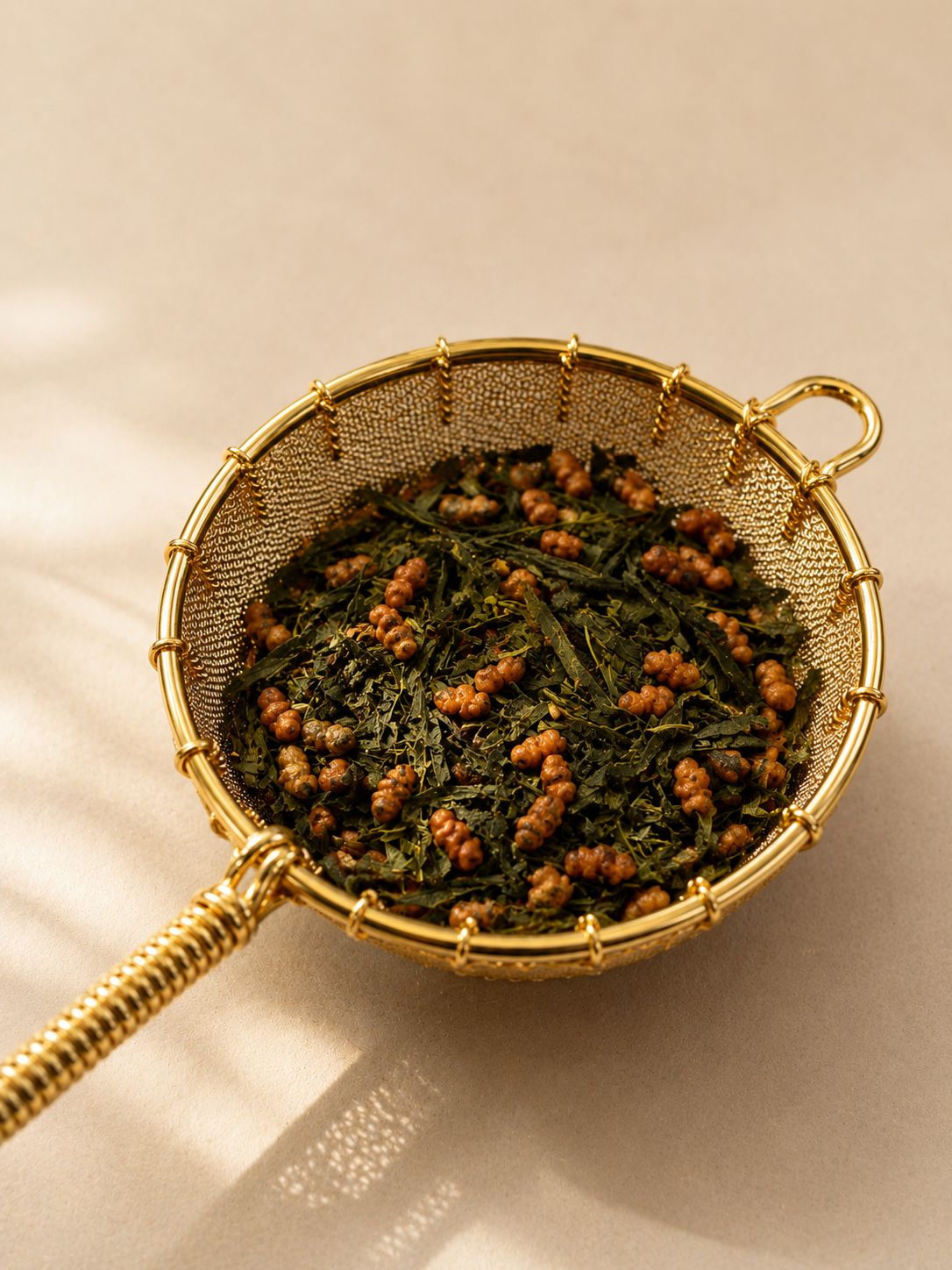 Close-up of a gold mesh tea strainer filled with genmaicha tea against a soft neutral beige background with warm natural shadows
