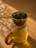Jasmine dragon pearls tea steeping in glass cup with gold strainer, light golden green tea infusion on wooden table
