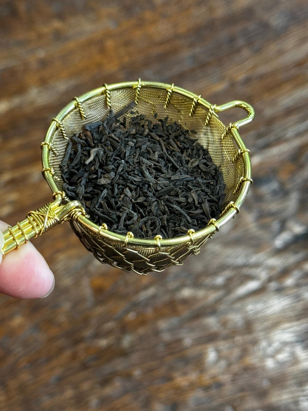 Premium organic Pu-erh loose leaf tea with dark twisted leaves displayed in a handcrafted brass strainer, ideal for traditional tea preparation.