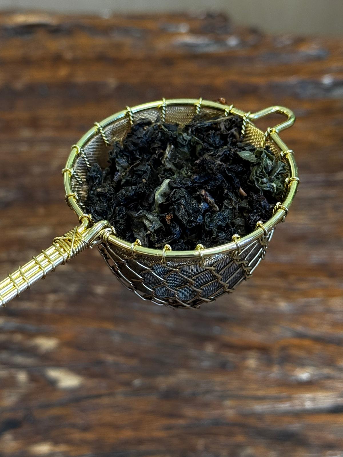 Organic Osmanthus Oolong loose leaf tea displayed in a handcrafted brass tea strainer, featuring dark rolled leaves with floral aroma and premium Taiwanese oolong character.