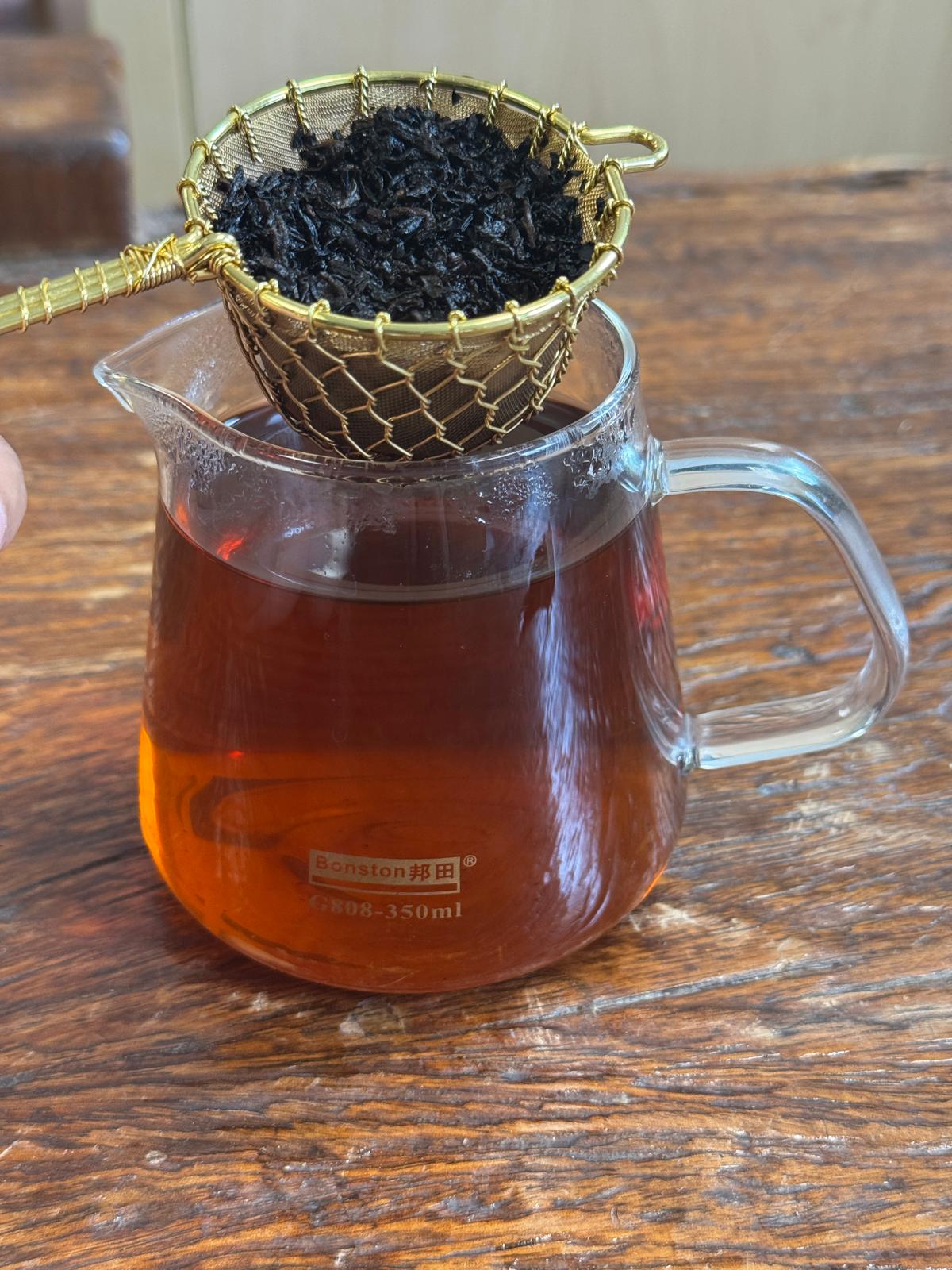 Steeped organic Pu-erh tea leaves suspended above a glass tea pitcher filled with dark amber tea, illustrating traditional loose leaf tea brewing.