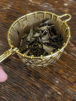 Organic Huang Shan White Tea leaves displayed in a gold mesh tea strainer on a rustic wooden table. Premium loose-leaf white tea from Yellow Mountain (Huang Shan), known for its delicate floral aroma, smooth taste, and antioxidant-rich properties.