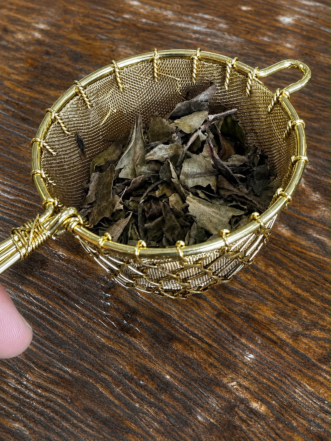 Organic Huang Shan White Tea leaves displayed in a gold mesh tea strainer on a rustic wooden table. Premium loose-leaf white tea from Yellow Mountain (Huang Shan), known for its delicate floral aroma, smooth taste, and antioxidant-rich properties.