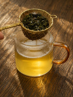 Glass teapot with tea leaves being poured into it on a wooden surface