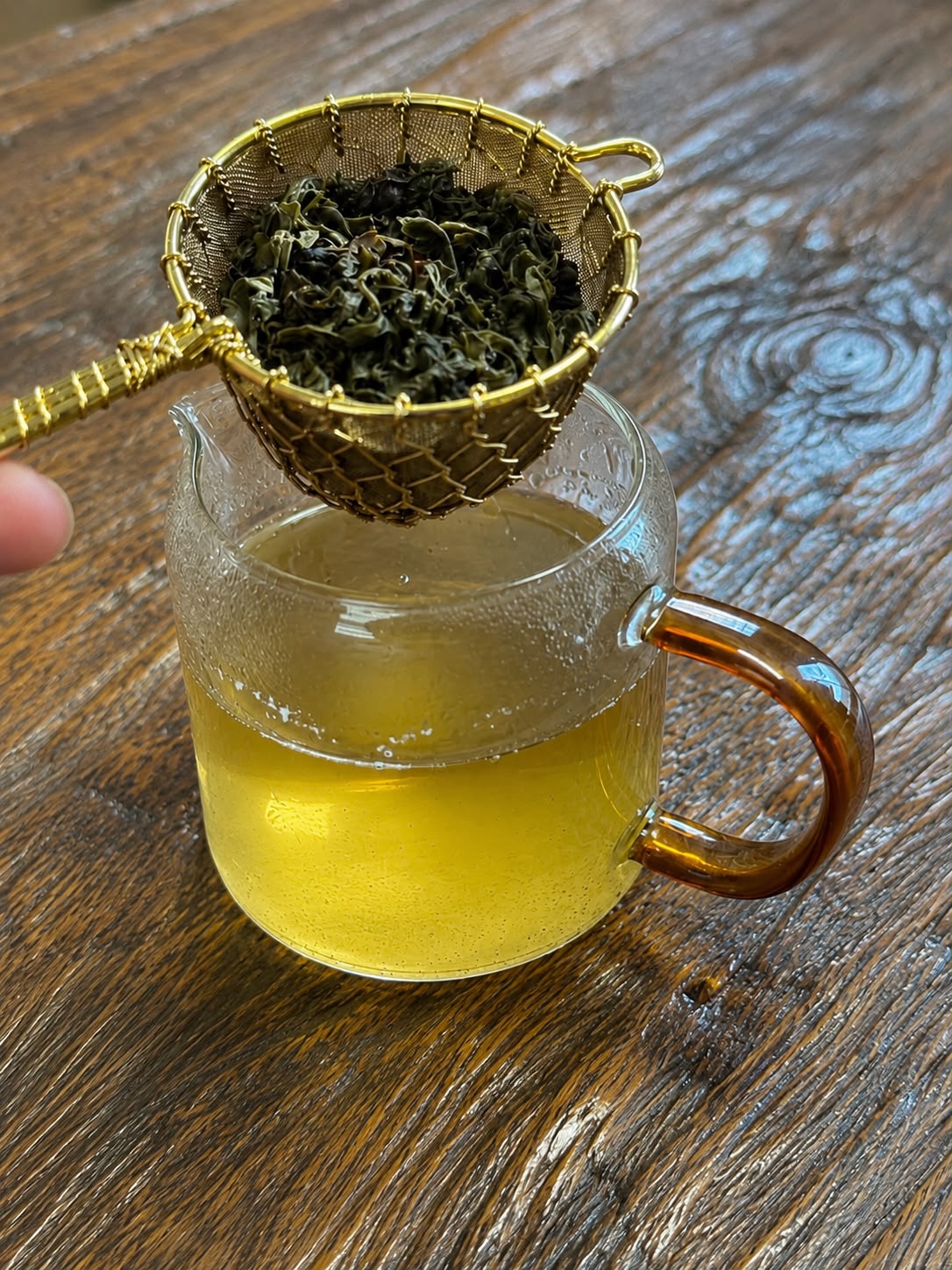 Tea strainer with green tea leaves over a glass of brewed tea on a wooden surface