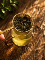 Organic Joonjak Tea loose leaves in a handcrafted gold tea strainer above a glass cup of freshly brewed green tea.
