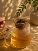 Hand holding handcrafted brass tea strainer with gold mesh, aesthetic loose leaf tea setup in warm natural light